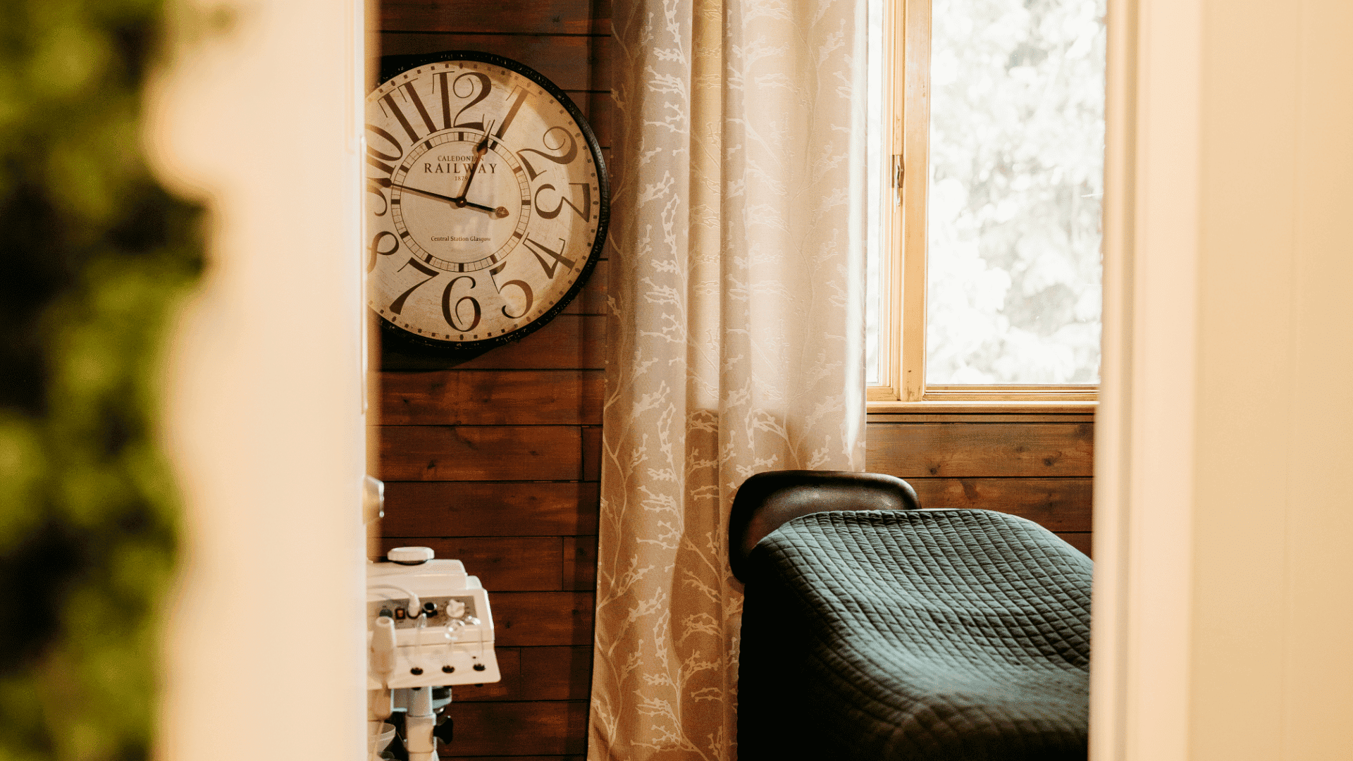 Cozy spa room with a massage table, vintage clock, and a window with sheer curtains.