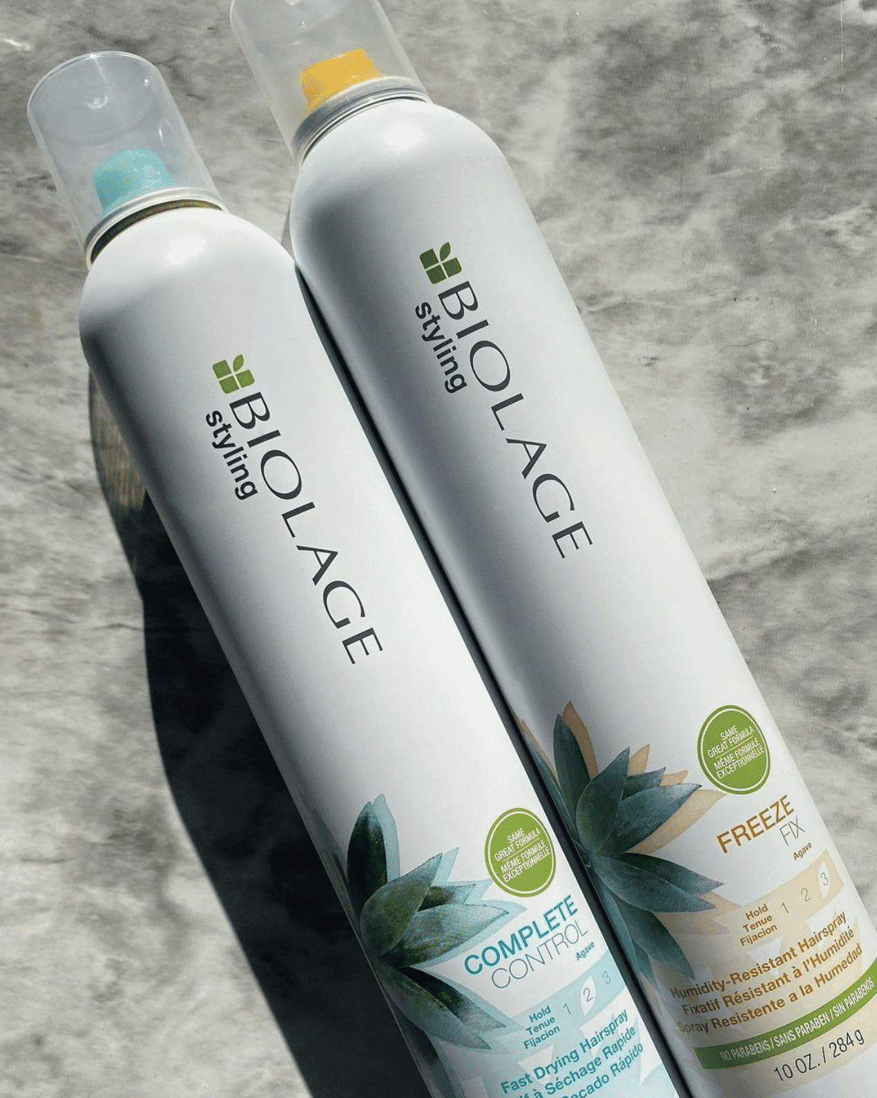 Two Biolage styling spray bottles on a marble surface, labeled 'Complete Control' and 'Freeze Fix.