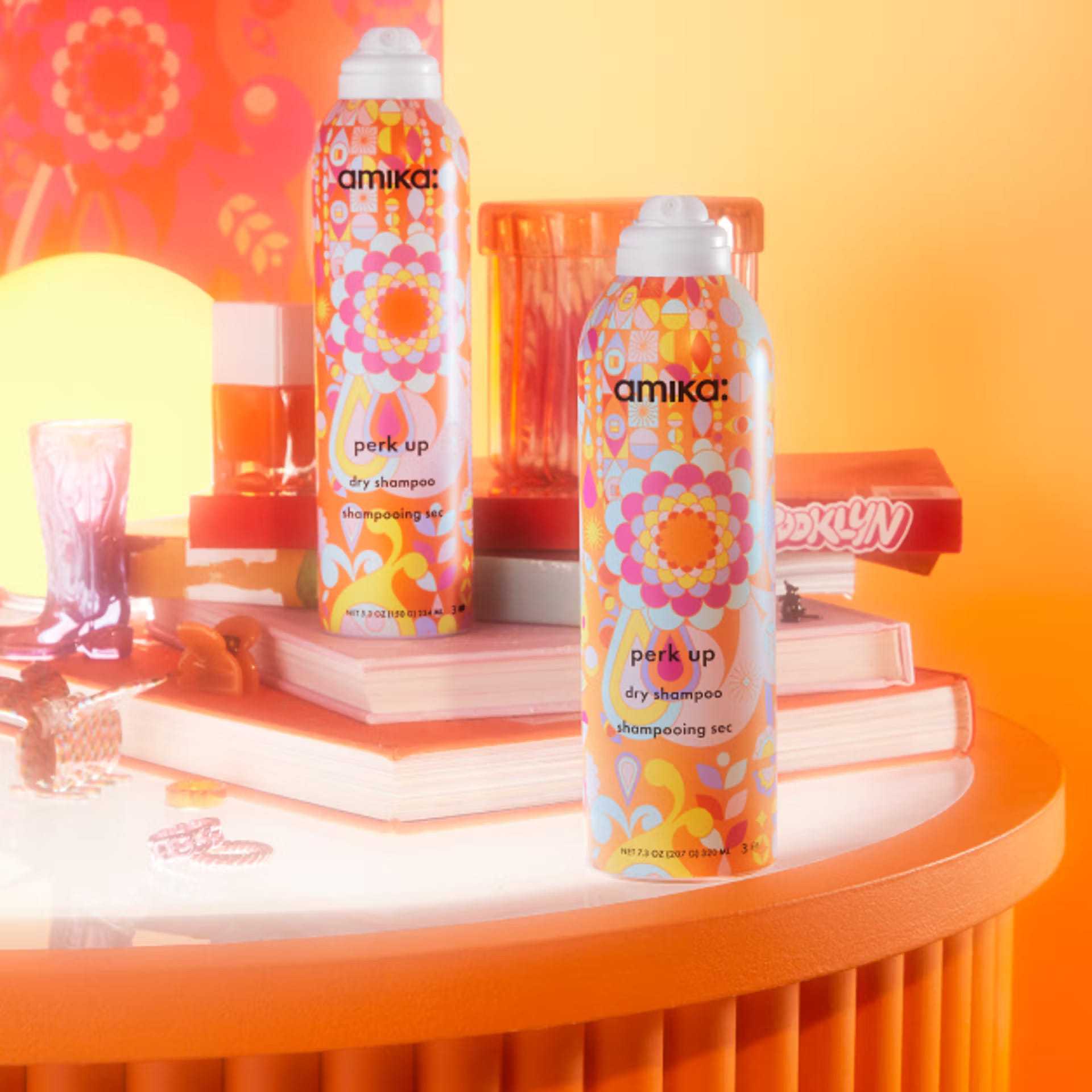 Two Amika Perk Up Dry Shampoo bottles on a vibrant, colorful vanity with books and decor.